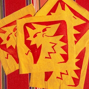 以下が含まれることがあります： 赤いスタイリッシュな星形のデザインが施された黄色の布の正方形が3つ。正方形は互いに積み重ねられており、赤と白のストライプの背景の上に置かれています。