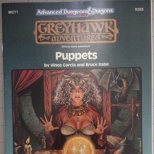 May include: A vintage "Greyhawk Adventures" game book cover. The cover features a fantasy illustration of a woman with curly hair, a crystal ball, and the title "Puppets." The book is from the Advanced Dungeons & Dragons series.