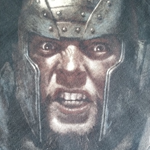 May include: A close-up of a man's face with a fierce expression. He is wearing a metal helmet with a textured surface. The helmet has a dark gray color and is decorated with rivets. The man's face is painted with a dark brown color and he has a thick beard. His eyes are wide open and his mouth is open in a shout.