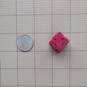 May include: A red, square-shaped rubber stamp with a cartoon skull design. The stamp is about the size of a quarter.