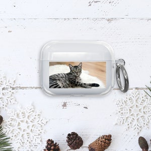Puede incluir: Una funda de tel&eacute;fono de pl&aacute;stico transparente con una foto de un gato atigrado impresa en la parte posterior. La funda tiene un anillo de metal plateado.