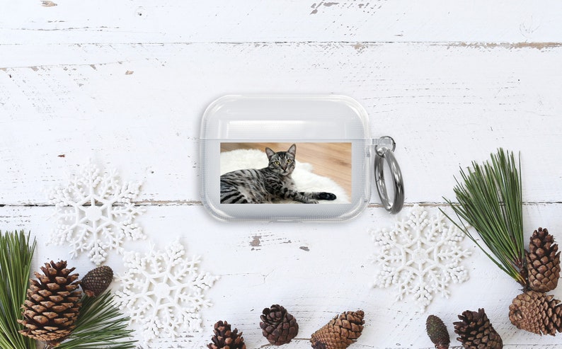 Puede incluir: Una funda de tel&eacute;fono de pl&aacute;stico transparente con una foto de un gato atigrado impresa en la parte posterior. La funda tiene un anillo de metal plateado.