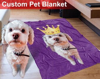 May include: A purple fleece blanket with a white dog wearing a gold crown printed on it.  The blanket is on a wooden floor.