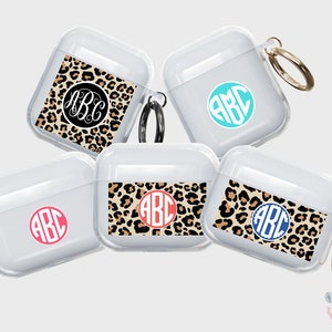 May include: Five clear AirPod cases with different designs.  One case has a black and tan leopard print with a white monogram "ABC". Another case has a turquoise monogram "ABC".  A third case has a coral monogram "ABC".  A fourth case has a navy blue monogram "ABC" on a black and tan leopard print.  The fifth case has a black and tan leopard print with a gold key chain.