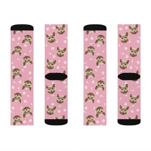 May include: Four pairs of pink socks with a repeating pattern of a cat's face and white paw prints. The socks have black cuffs.