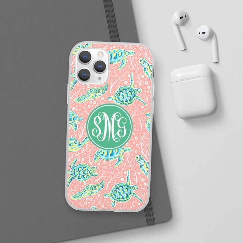 May include: A clear phone case with a pink background and a pattern of green and blue sea turtles. The case has a green circle with the monogram "SMC" in white.