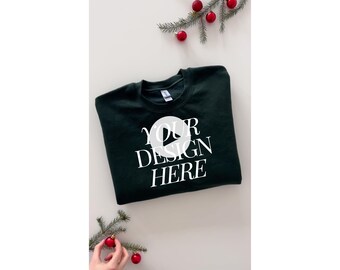 May include: A dark green sweatshirt with the text "YOUR DESIGN HERE" in white on the front. The sweatshirt is folded and laid flat on a white surface. There are red Christmas ornaments and green pine branches on the surface.