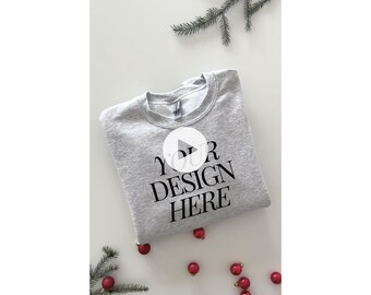 May include: A gray sweatshirt with the text "YOUR DESIGN HERE" printed on the front. The sweatshirt is folded and lying on a white surface with red ornaments scattered around it.