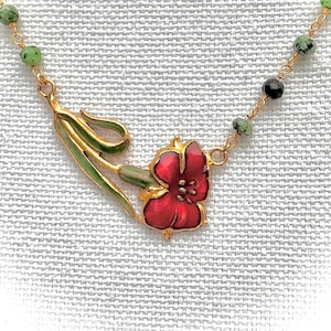 May include: A gold-toned necklace featuring a red flower pendant with green and gold accents. The necklace has a delicate chain with green and black beads. The flower pendant is detailed with red enamel and green leaves.