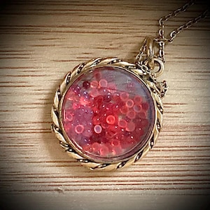 Peut inclure: Un collier avec un pendentif en or et un médaillon rond en verre rempli de paillettes rouges. Le médaillon est orné d'une bordure en corde torsadée.