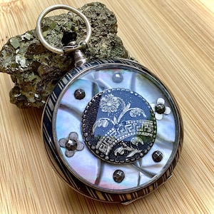 Könnte beinhalten: Eine silberne Taschenuhr mit einem Perlmutt-Zifferblatt und einem schwarzen Emaille-Blumendesign. Die Uhr hat eine silberne Kette und ist mit schwarzen und weißen Streifen verziert.