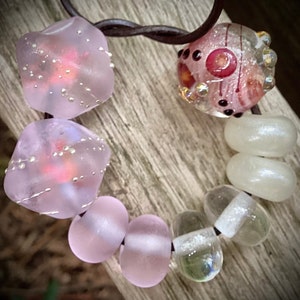 Puede incluir: Un primer plano de un collar de cuentas de vidrio rosa y blanco. El collar presenta una variedad de formas y tamaños de cuentas, incluyendo redondas, cuadradas y ovaladas. Las cuentas son todas translúcidas y tienen un acabado brillante.