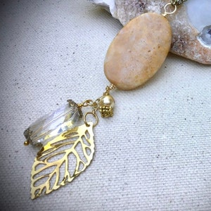 Op de afbeelding: Een gouden ketting met een grote, ovale hanger van lichtbruine steen. De hanger is bevestigd aan een ketting met een heldere kristallen kraal, een parelkraal en een gouden bladbedel.