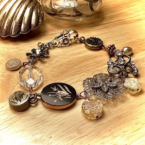 May include: A vintage-inspired bracelet featuring a mix of metal, glass, and crystal beads. The bracelet includes a variety of charms, such as a flower, a button, and a crystal. The bracelet is crafted with copper wire and has a silver clasp.