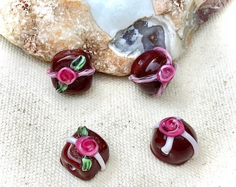 May include: Four glass beads shaped like gift boxes with pink roses and white ribbon accents. The beads are a dark brown color.