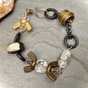 Peut inclure: Un bracelet fait à la main avec un design unique. Il comprend des éléments métalliques couleur laiton, des perles facettées transparentes, des anneaux en caoutchouc noirs et un fermoir barillet couleur laiton. Le bracelet a une esthétique d'inspiration industrielle.