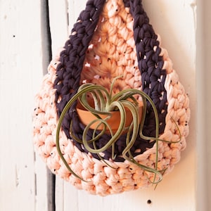 May include: A hanging macrame plant holder in peach, dark purple, and light pink. It contains a small terracotta pot with a green air plant. The macrame is suspended from a loop at the top. The plant holder is handmade.
