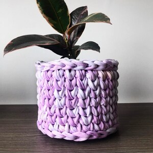 May include: A houseplant with dark green and burgundy leaves in a handmade, cylindrical basket. The basket is made from a thick, woven fabric in shades of lavender and white, creating a textured look. The plant is on a dark wooden surface.