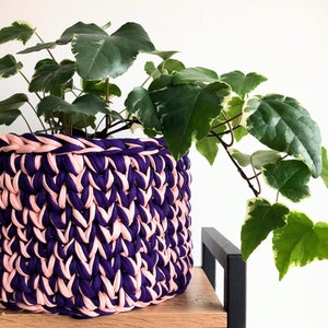 May include: A handcrafted crochet basket in shades of purple and pale pink, containing a green plant with variegated foliage. The basket is made from chunky yarn and has a textured finish. The plant pot is placed on a wooden shelf.