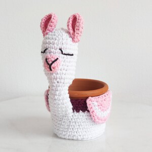 Peut inclure: Un pot de plante en crochet blanc en forme de lama, avec des détails roses sur les oreilles et le nez. Il contient un petit pot en terre cuite marron. Le pot fait main est idéal pour les petites plantes.