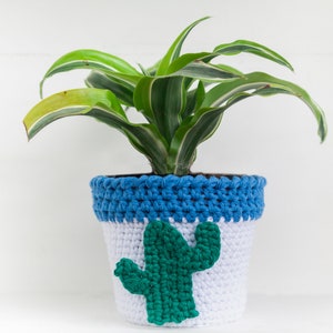 Peut inclure: Un pot de plante crocheté blanc et bleu avec un motif de cactus vert. Il contient une plante verte et blanche. Le haut du pot est bleu et le cactus est centré sur le devant. Le fond est blanc.