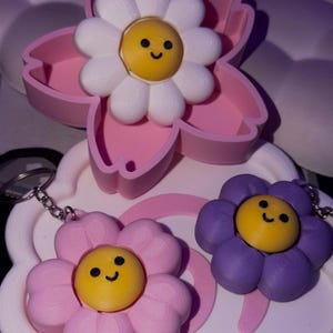 May include: A collection of flower-shaped keychains in pink, purple, and white. Each keychain features a smiling yellow center and a chain for attachment. One flower is inside a pink flower-shaped container.