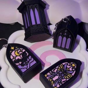 May include: A collection of black gothic-style keychains and a lantern. The keychains feature stained glass designs with purple, pink, and yellow accents. The lantern has a purple interior. The items are displayed on a white scalloped tray.