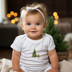 Könnte beinhalten: Ein Baby in einem weißen Body mit einer Aquarellillustration eines Weihnachtsbaums und einer Maus. Das Baby trägt ein weißes Stirnband mit einer Schleife und lächelt. Ein kleiner Weihnachtsbaum ist im Hintergrund.