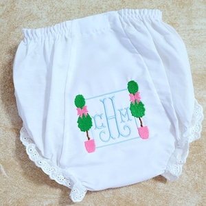 May include: White baby bloomers with a blue and green embroidered monogram and topiary design. The monogram is "CM" with a bow on each topiary.