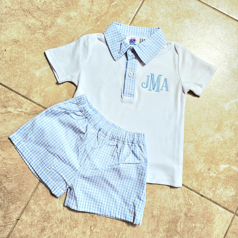 May include: A white polo shirt and matching shorts set. The shirt has a blue plaid collar and the embroidered initials "JMA" in blue. The shorts are blue plaid. The set is designed for children.