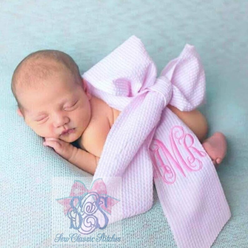 Pode incluir: Um envolt&oacute;rio de la&ccedil;o de gingham rosa e branco com um monograma rosa "MVR" &eacute; usado como um envolt&oacute;rio para um rec&eacute;m-nascido. O beb&ecirc; est&aacute; dormindo pacificamente.