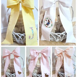 May include: A collage of decorative bows in yellow, white, and pink, each with a long streamer. Some bows have embroidered details like a floral initial, a bunny, or a wreath. The bows are placed in white baskets.