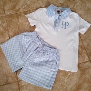 May include: A white polo shirt and matching shorts set. The shirt has a light blue checkered collar and the initials "JP" embroidered in light blue. The shorts are light blue and white checkered.