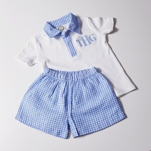May include: A white polo shirt and blue gingham shorts set. The shirt has a blue gingham collar and placket, with embroidered initials "THG" in blue. The shorts have an elastic waistband. This outfit is perfect for a baby or toddler.