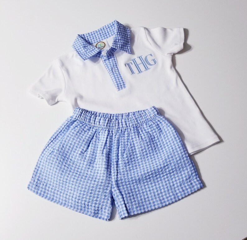 May include: A white polo shirt and blue gingham shorts set. The shirt has a blue gingham collar and placket, with embroidered initials "THG" in blue. The shorts have an elastic waistband. This outfit is perfect for a baby or toddler.