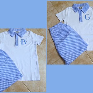 May include: Two white polo shirts with blue gingham collars and matching blue gingham shorts. Each shirt has a single initial embroidered in blue on the chest. The background is a light blue.