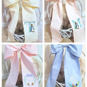 May include: A collage of four decorative bows in pastel colors: cream, pink, and blue. Each bow features a long, embroidered ribbon with a Peter Rabbit design and a letter or monogram. The bows are tied on white wicker baskets.
