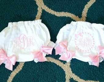 May include: Two white baby bloomers with pink embroidered monograms and pink satin bows. The bloomers have a frilled waist and lace trim on the legs. The background is a patterned green rug.