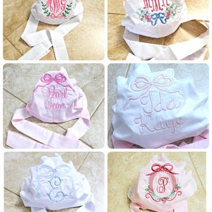 May include: Four white fabric bonnets with pink and blue ribbon ties. Each bonnet is embroidered with a different design, including a monogram, a name, and a bow. The bonnets are displayed on a tiled surface.