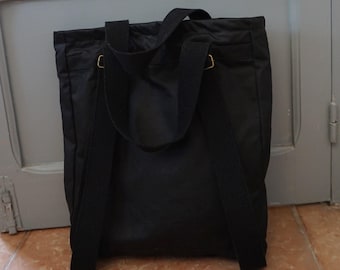 May include: A black tote bag featuring two wide fabric shoulder straps and two additional vertical straps that extend down the front. The bag includes small gold-toned metal hardware rings where the straps attach to the main body of the bag.