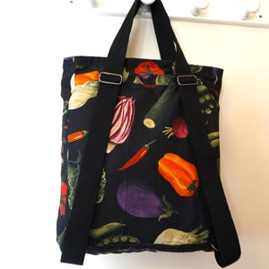 May include: A black fabric backpack featuring a colorful vegetable print with illustrations of orange bell peppers, purple eggplants, red chili peppers, and green zucchini. The bag includes two black adjustable shoulder straps and metal hardware.