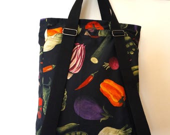May include: A black fabric backpack featuring a colorful vegetable print with illustrations of orange bell peppers, purple eggplants, red chili peppers, and green zucchini. The bag includes two black adjustable shoulder straps and metal hardware.