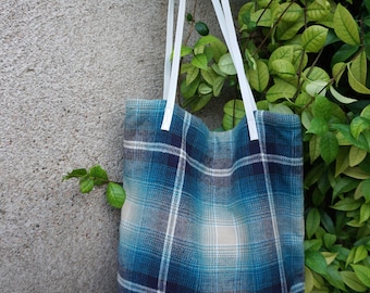 May include: A blue plaid tote bag with white straps hangs against a textured gray wall, with green foliage in the background. The bag features a pattern of blue, teal, and white squares.