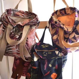 Peut inclure: Trois sacs fourre-tout avec poignées supérieures et fermetures éclair. Un sac présente un motif de feuilles rouge-brun, un autre un design géométrique de pétales violets et beiges, et le troisième est noir avec un imprimé de légumes colorés.
