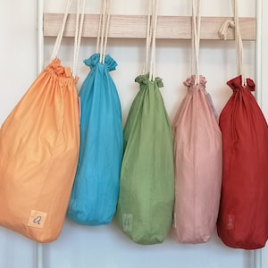 Op de afbeelding: Vijf trekkoordtassen in verschillende kleuren hangen aan een wit rek. De tassen zijn oranje, blauw, groen, roze en rood. Elke tas heeft een klein label met de letter "a" erop.