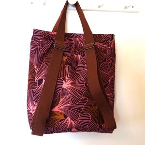 Könnte beinhalten: Ein wandelbarer Rucksack-Shopper aus dunkelviolettem Stoff mit einem sich wiederholenden Ginkgo-Blattmuster in Rosa-, Orange- und Dunkelrottönen. Die Tasche verfügt über zwei braune Webgurte mit verstellbaren Metallschnallen zum Tragen.