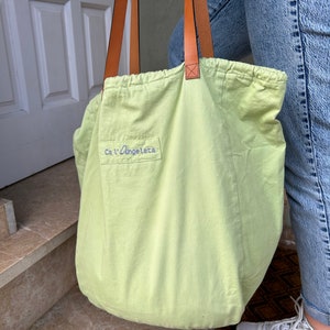 May include: A light green canvas tote bag with brown leather straps. The bag has a drawstring closure and the text "Ca'l'Angeletta" is embroidered on the front.