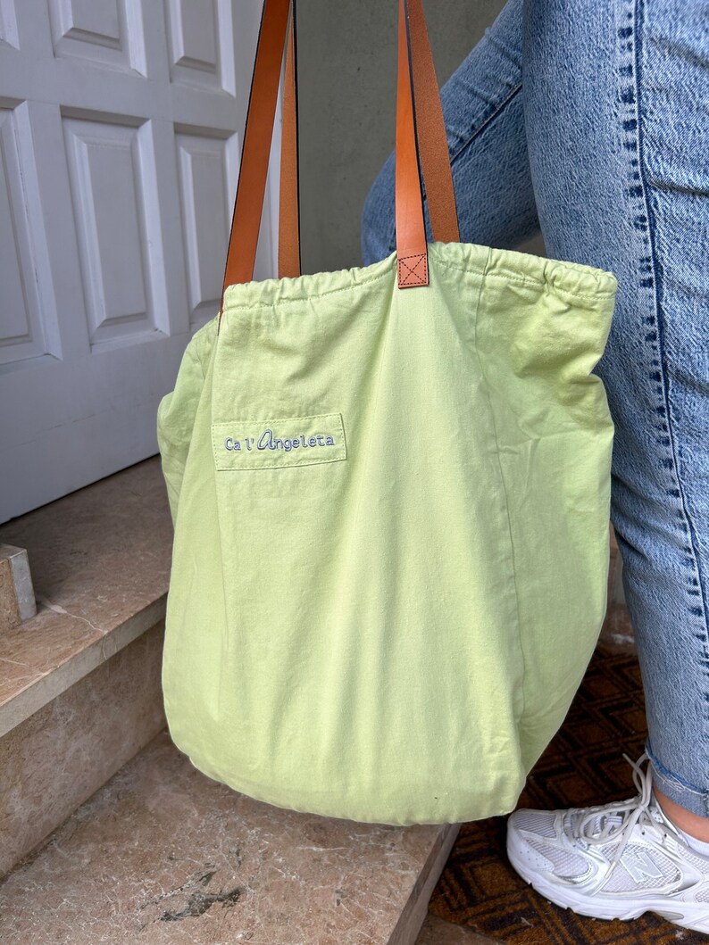 May include: A light green canvas tote bag with brown leather straps. The bag has a drawstring closure and the text "Ca'l'Angeletta" is embroidered on the front.