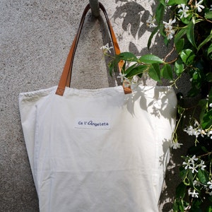 May include: A white canvas tote bag with brown leather straps and a blue label that says "Ca l'Angeleta".
