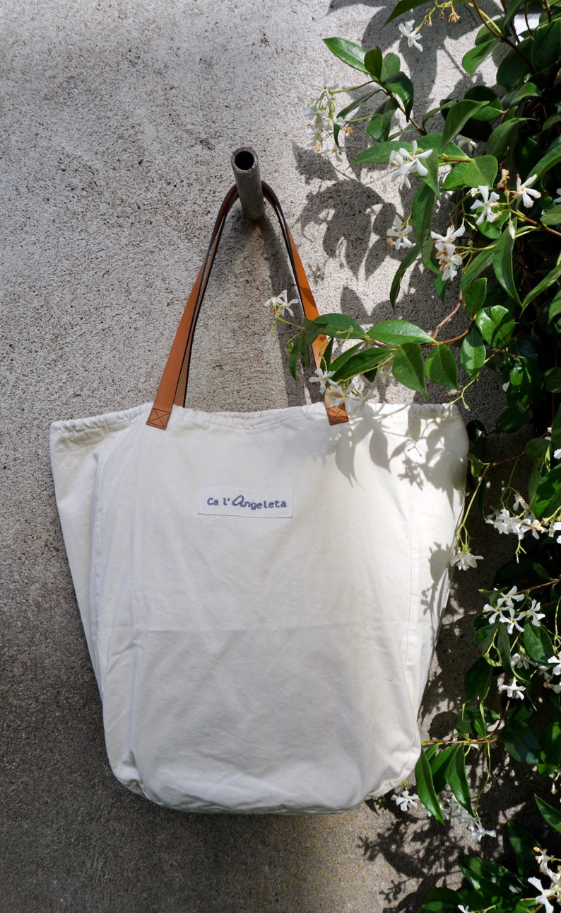 May include: A white canvas tote bag with brown leather straps and a blue label that says "Ca l'Angeleta".